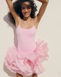 Smiling model with dark curly hair wearing a pink ruffled dress against a plain neutral background.