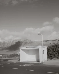 Black and white photograph of an empty white concrete bus stop shelter and lamppost on a roadside with mountains in the background.