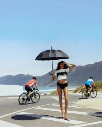 Model holding a black umbrella on a coastal road with two cyclists riding in the background under a clear sky.