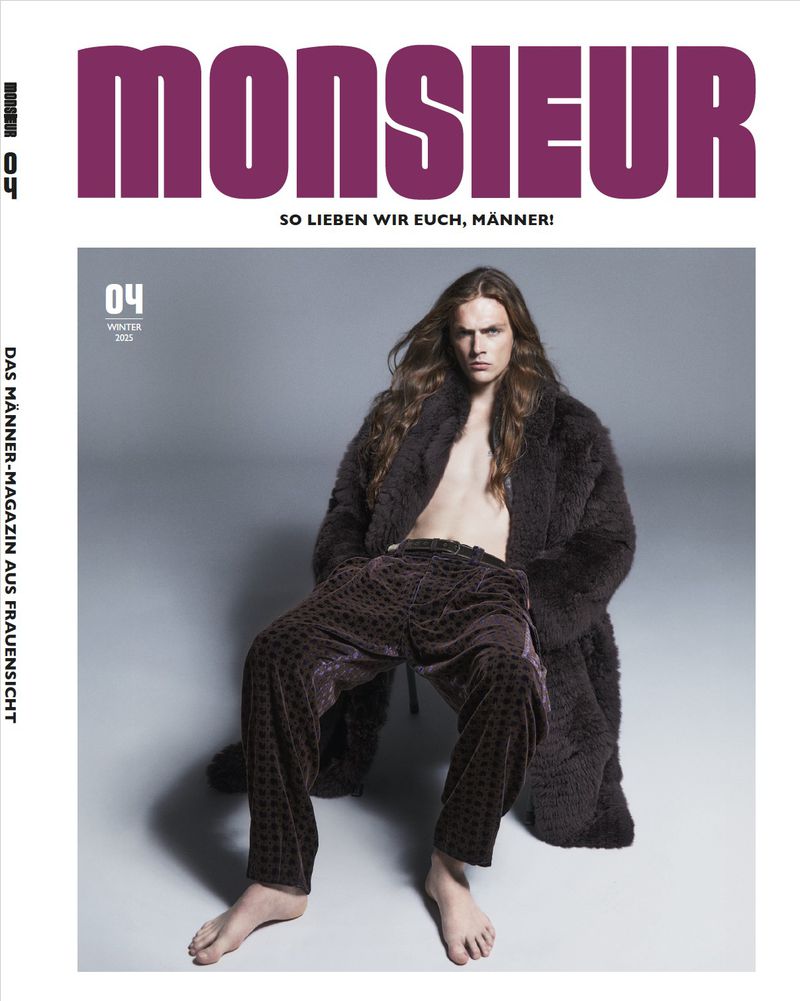 Magazine cover featuring a bare-chested male model with long brown hair, wearing patterned pants and a dark fur coat, seated against a grey background.