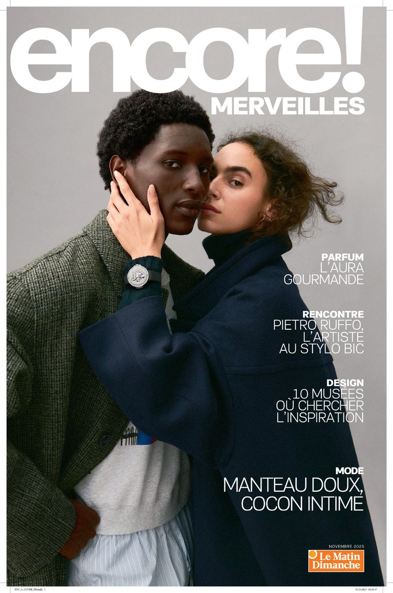 Magazine cover featuring a dark-skinned male and a light-skinned female in coats, embracing with a soft, neutral-toned backdrop and elegant text.