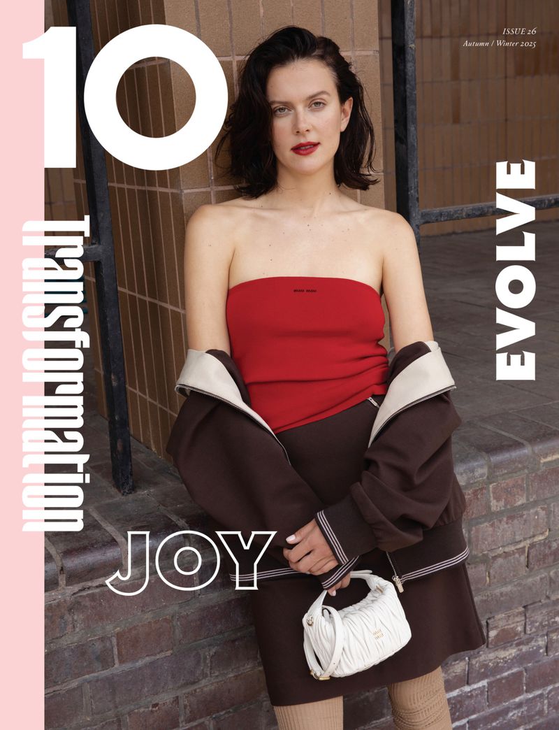 A young adult woman poses in a red top and brown skirt, holding a white purse, next to a brick wall. Magazine cover aesthetic.