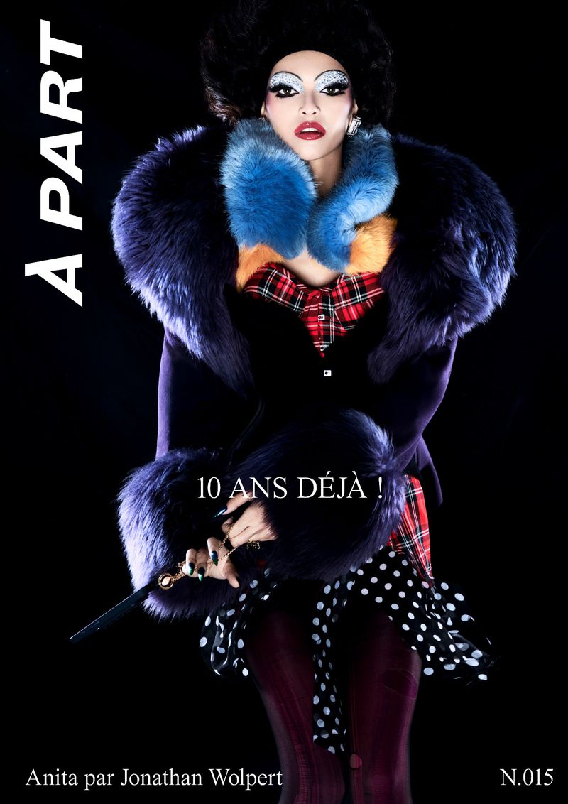Elegant person with heavy makeup, fur-trimmed coat, holding a cane, posing against a black backdrop for a studio fashion shot.