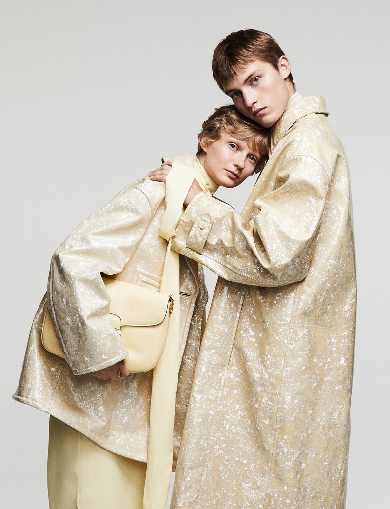Two fashion models wearing matching patterned trench coats in a studio setting, one holding a pale yellow shoulder bag with a tie scarf.