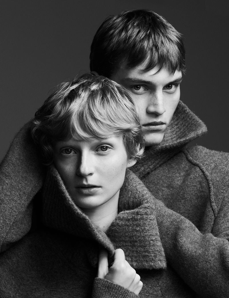 Monochrome portrait of a young adult couple in wool sweaters. The woman is in front, with the man behind her, both looking at the camera.