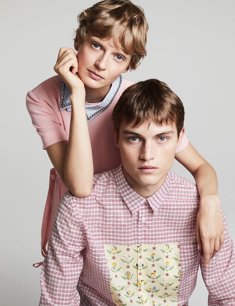 Two fair-skinned people pose in a studio setting. The person in the foreground wears a patterned pink button-down shirt. The person in the background wears a pink top.