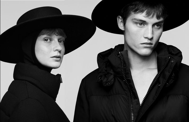 Two people model fashionable black puffer jackets and wide-brimmed hats against a light background in a studio setting, portrait orientation.