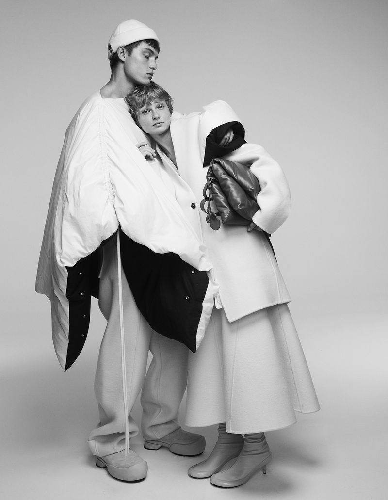 Monochrome fashion shot of two young adult people in white coats and beige outfits posing together in a light studio setting with modern styling.