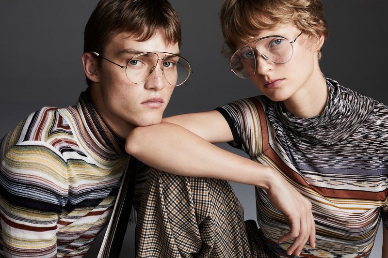 Two stylish people wearing glasses and patterned clothing are posing indoors against a neutral background in a fashion forward portrait.