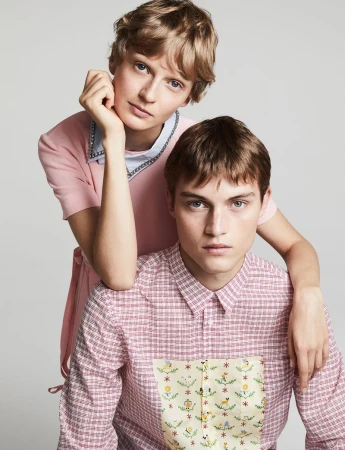 Two models posing together against a plain grey backdrop, a woman in a pink dress resting her hand on a man in a patterned shirt.