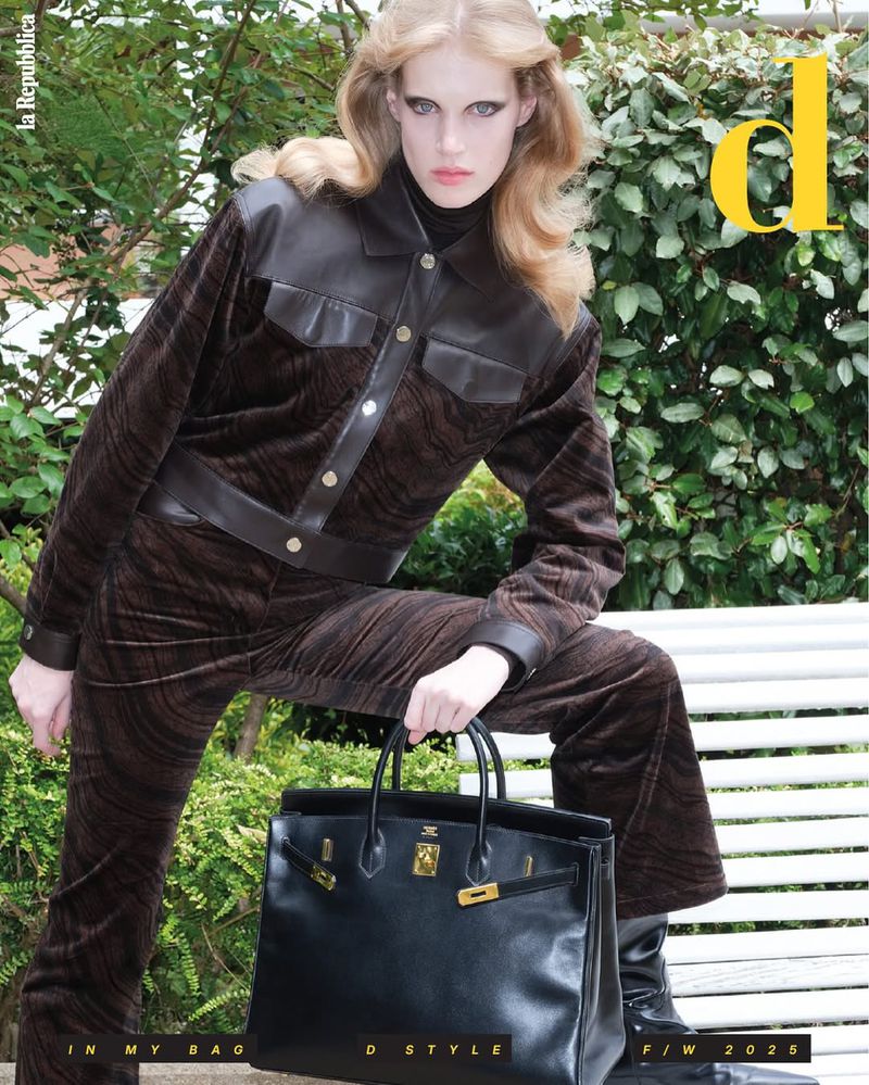 Blonde model poses with black handbag in brown jacket and pants on a white bench outdoors against a green leafy background for a fashion editorial.