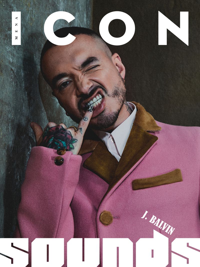 Magazine cover featuring J. Balvin with a pink blazer, winking, and holding a finger to his mouth, set against a textured wall.