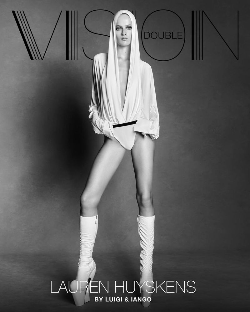 A fashion shot of a model in a white bodysuit, long white boots, and a draped white hooded top against a neutral gray background.