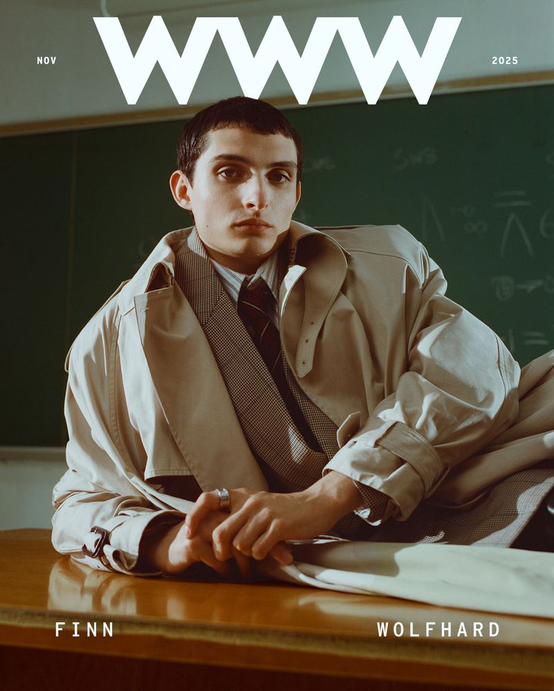 Magazine cover featuring a young adult male model in a trench coat, shirt, and tie, posing on a wooden desk in front of a chalkboard.