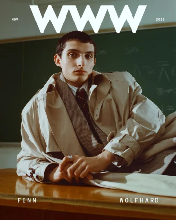 Magazine cover featuring a young adult male model in a trench coat, shirt, and tie, posing on a wooden desk in front of a chalkboard.