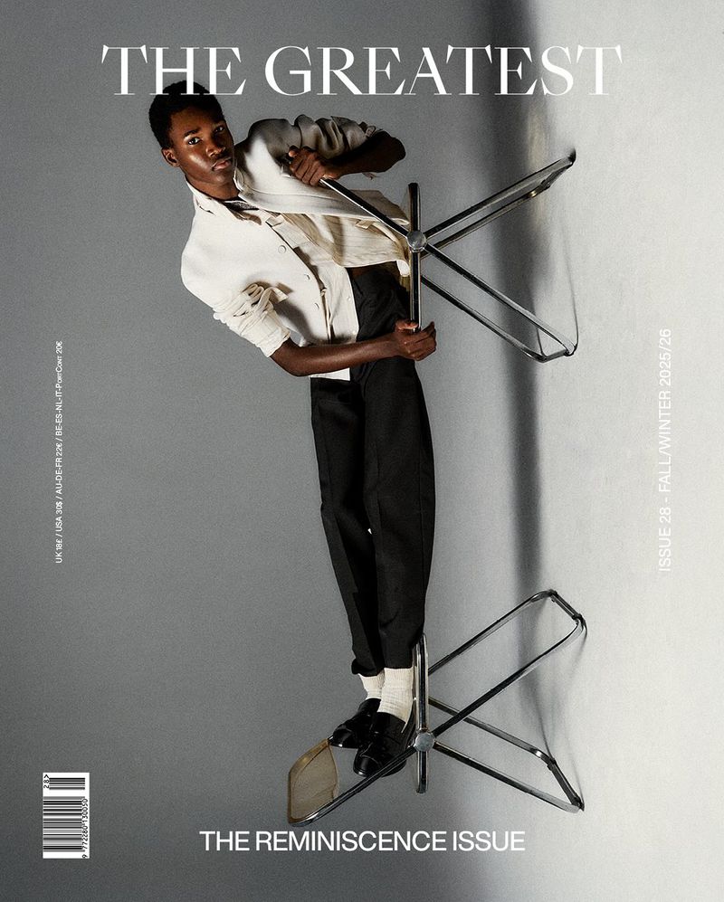 Fashion editorial shot of a Black male model in a white jacket and black pants balancing on two chairs against a gray background.