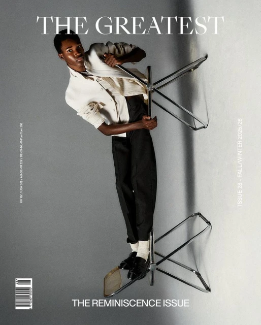 Fashion editorial shot of a Black male model in a white jacket and black pants balancing on two chairs against a gray background.