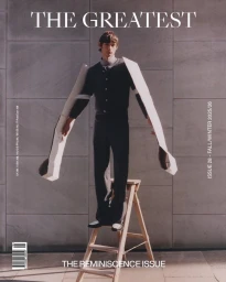 Male model standing on a wooden ladder wearing an outfit with elongated sculptural cardboard sleeve extensions against a stone wall.