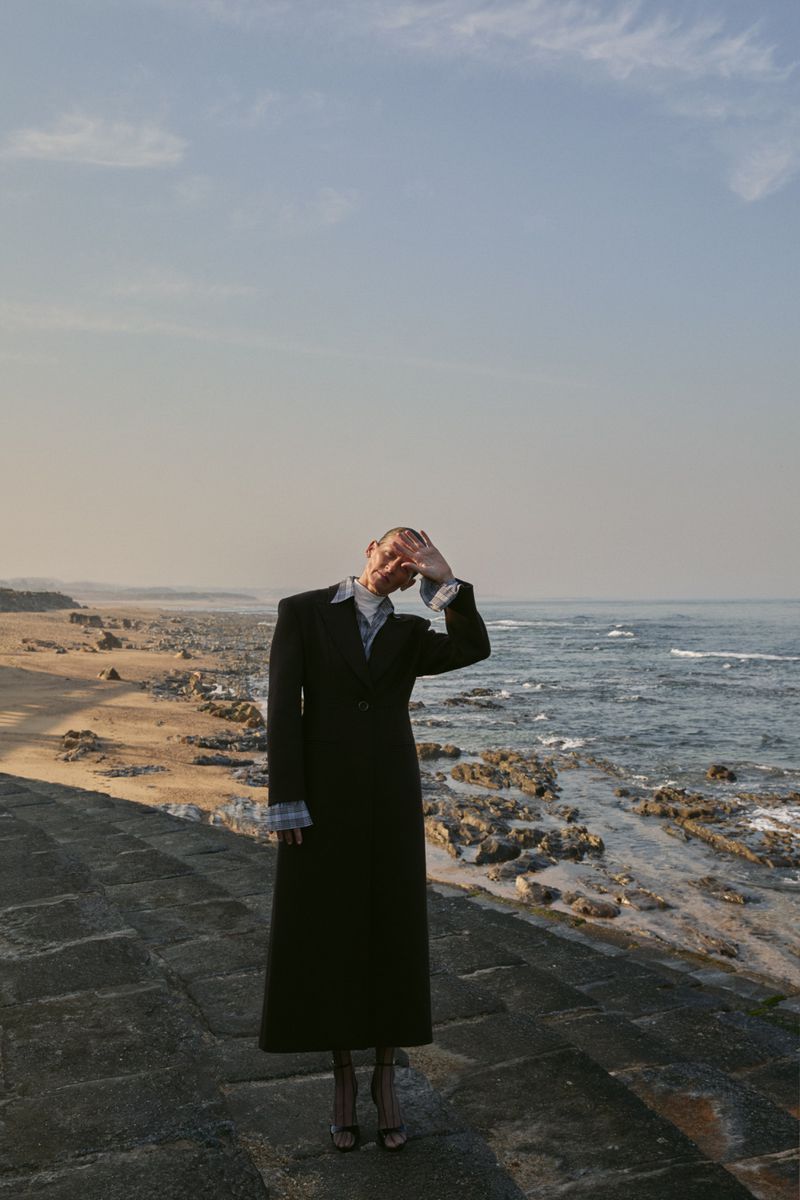 A person in a dark coat stands on a concrete surface near the ocean, shielding their eyes from the sun in an outdoor fashion shot.