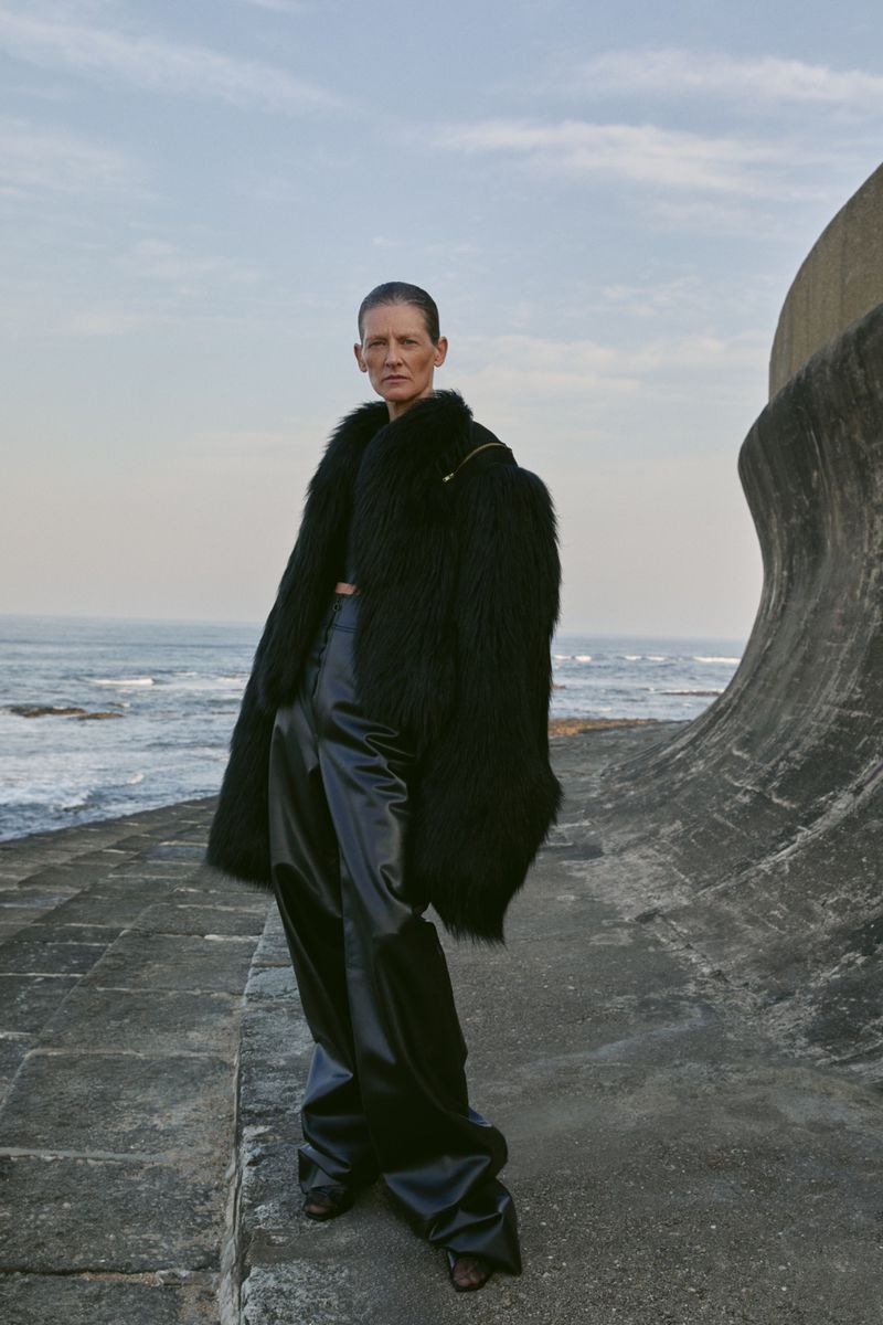 Fashion shot of an adult female wearing a black fur coat and shiny black pants, posing on a concrete platform by the ocean on a cloudy day.