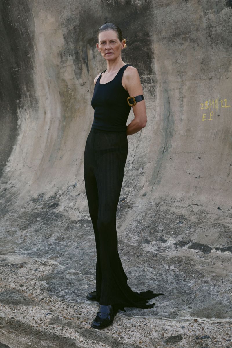 Woman in black tank top and skirt poses in front of weathered concrete wall with soft natural lighting, fashion editorial style.
