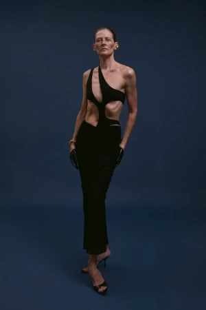 Elegant woman poses in a stylish black cutout dress, gold bracelet, earrings, and black heels against a solid dark blue background.