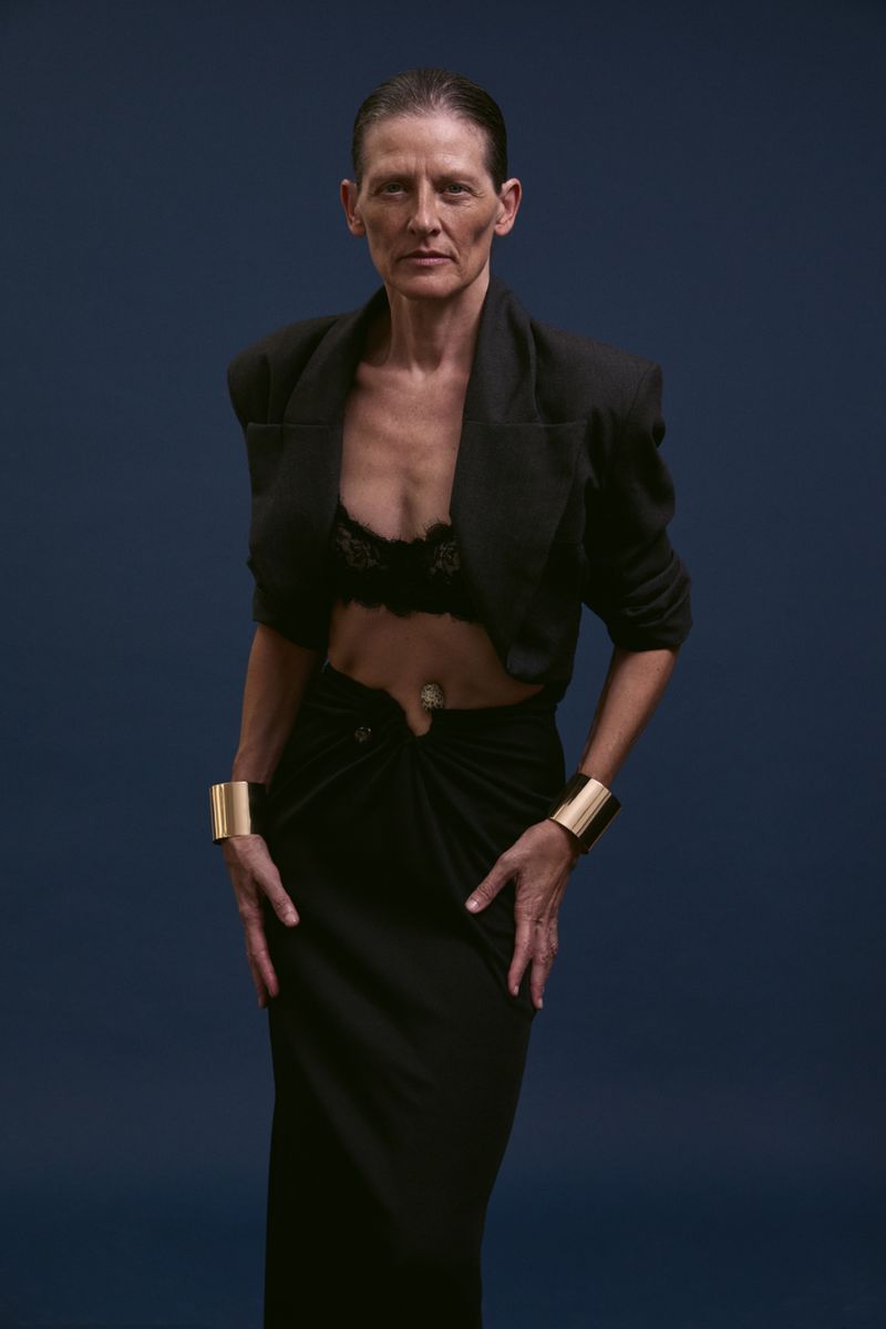 Elegant woman in a black cropped blazer and matching long skirt poses against a dark blue backdrop, showcasing fashion jewelry.
