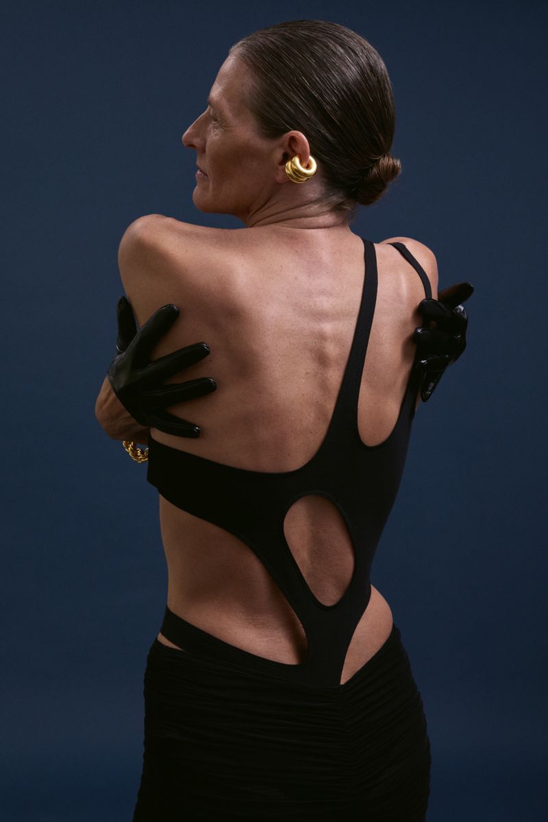 Elegant older woman with back to camera in cutout black dress and black gloves, accessorized with gold earrings against navy background.