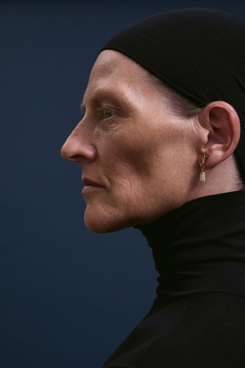 Profile view of an adult female with fair skin, wearing a black headscarf and turtleneck against a dark blue background in a studio.