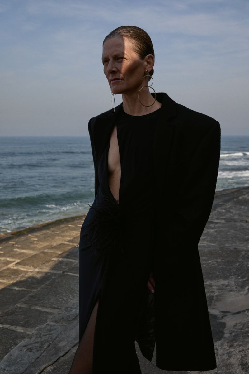 A woman poses on a stone ledge with the sea in the background, wearing a black dress with large circular earrings, under natural light.