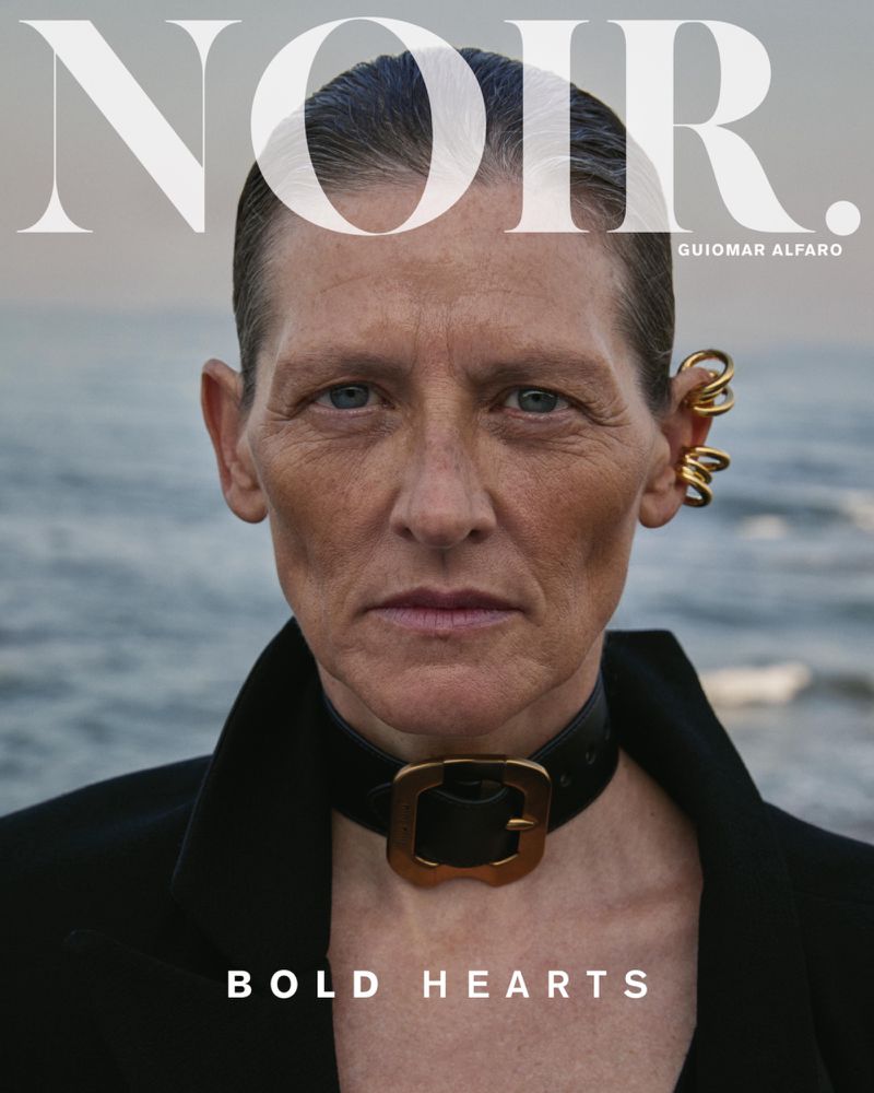 Close-up magazine cover shot of an adult woman with short hair wearing a black jacket and gold statement choker necklace near the ocean.