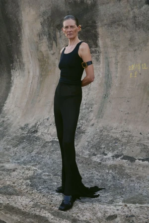 Model standing against a weathered concrete wall wearing a black top and black trousers for Noir Magazine editorial
