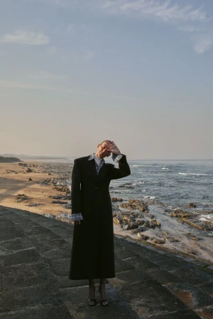 Model Guiomar Alfaro in a long black tailored coat standing on stone steps by a rocky coastline, shielding her eyes.