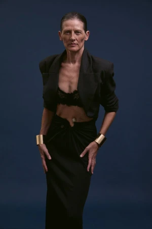 Model Guiomar Alfaro posing in a black cropped blazer, black skirt, and gold cuff bracelets against a dark blue background.