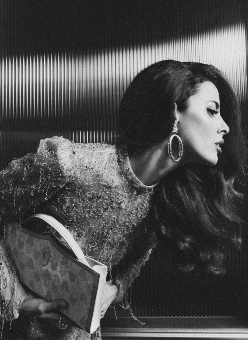 Elegant woman in ornate dress holds patterned purse in a black-and-white fashion shot, accented by large earrings and retro hairstyle.