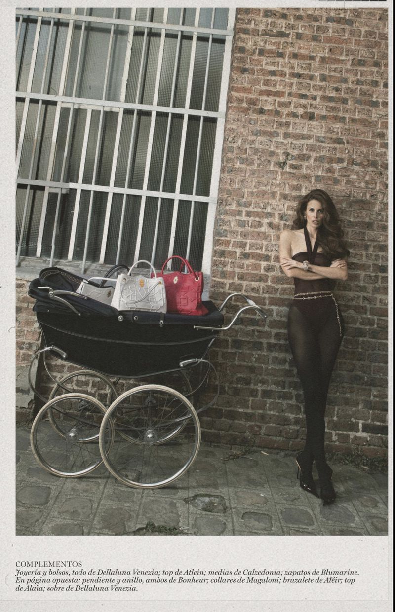 Fashion editorial shot of a woman in tights standing near a baby stroller filled with handbags in front of a brick wall with a window.