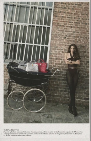 Fashion editorial shot of a woman in tights standing near a baby stroller filled with handbags in front of a brick wall with a window.