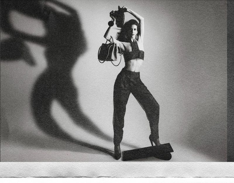 Monochrome fashion portrait of a woman in a cropped top and trousers holding a handbag, casting a dramatic shadow against a studio wall.