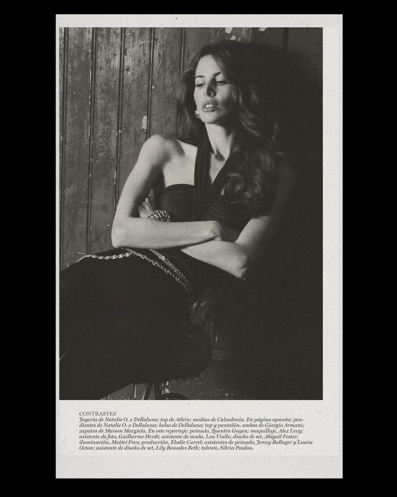 Model with long dark wavy hair in a black halter top and silver chain belt leaning against a rustic dark wooden wall.