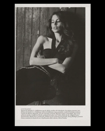 Model with long dark wavy hair in a black halter top and silver chain belt leaning against a rustic dark wooden wall.