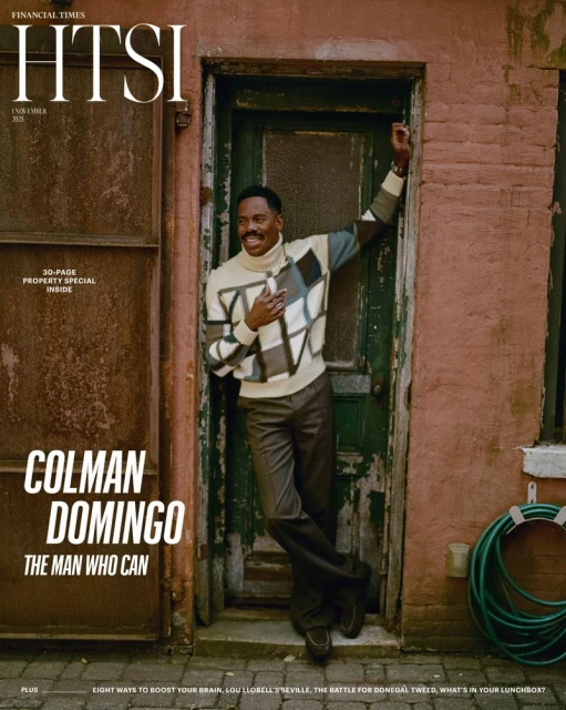 Colman Domingo poses for a magazine cover. He is wearing a patterned sweater, standing against a weathered brick wall, smiling at the camera.