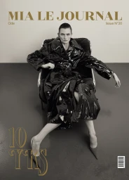 A model seated in a dark armchair wearing a glossy black trench coat against a neutral studio background for Mia Le Journal