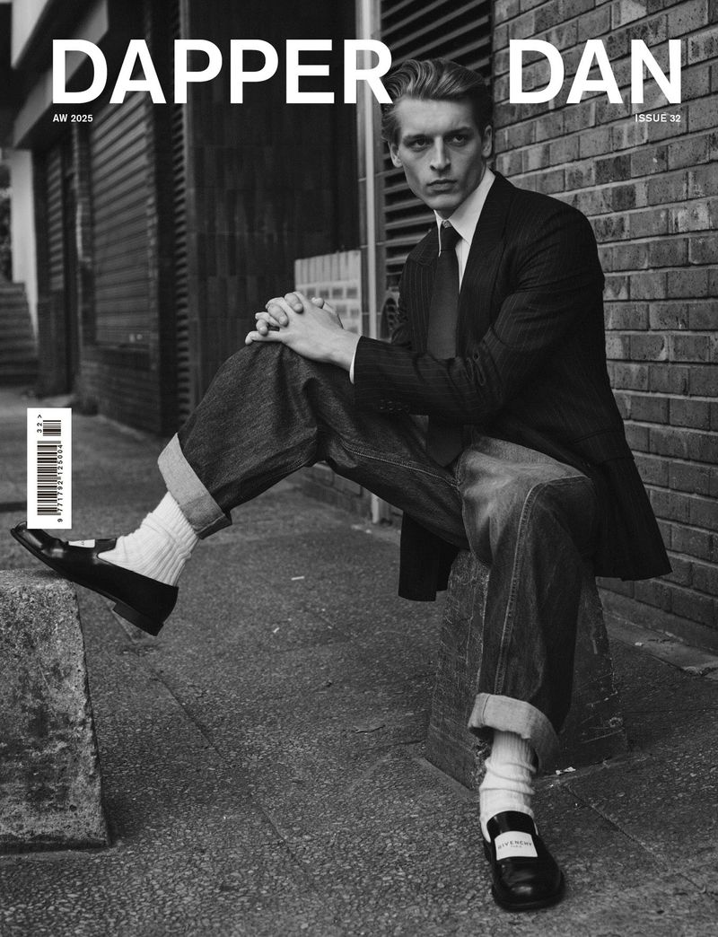 Model wearing a dark blazer, necktie, wide cuffed jeans, and loafers seated against a brick wall for Dapper Dan magazine.