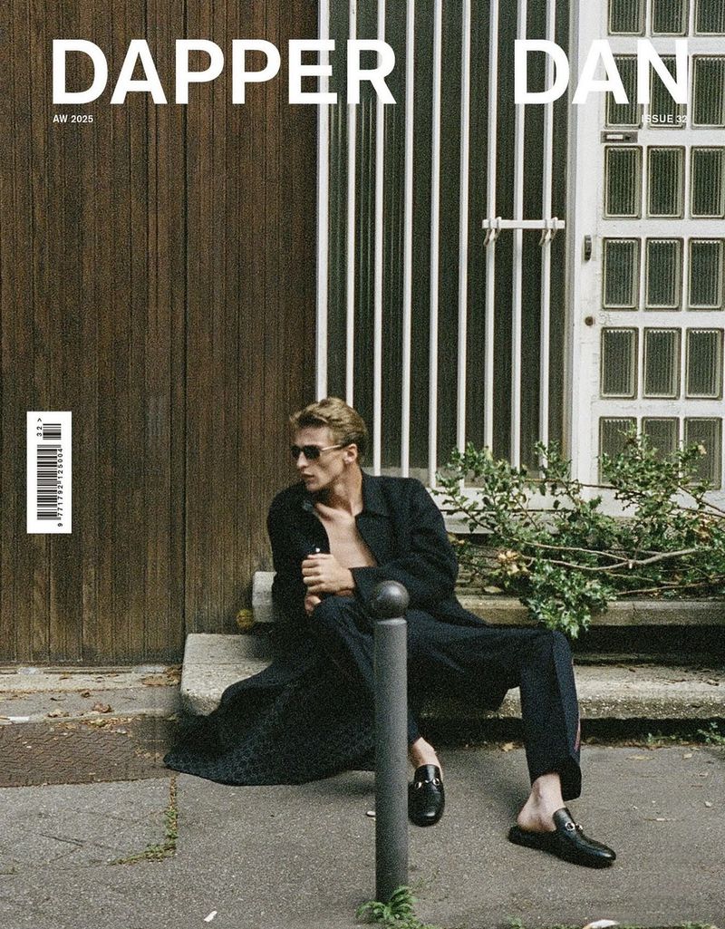 Fashion editorial: Man poses in sunglasses and a coat, sitting on concrete steps with a white door and wood wall backdrop outdoors.