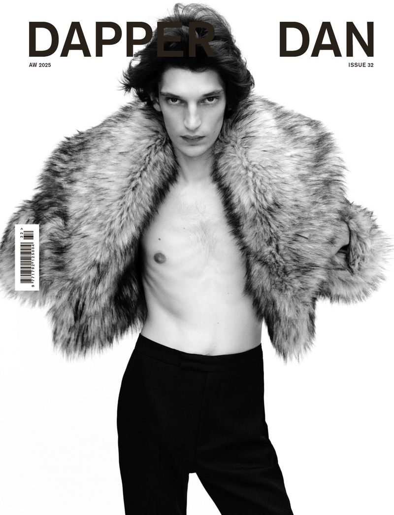 Black and white portrait of a slender adult male fashion model with dark hair, wearing a fur coat and standing against a white backdrop.