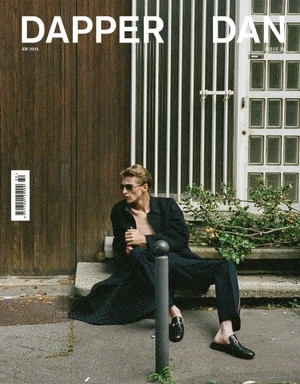 A man wearing sunglasses and an open black coat seated on outdoor steps for Dapper Dan Magazine issue 32.