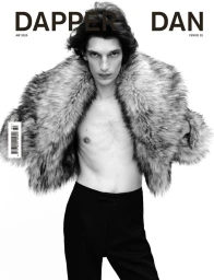 A male model posing shirtless wearing a voluminous fluffy fur jacket and black trousers against a white background for a magazine cover.