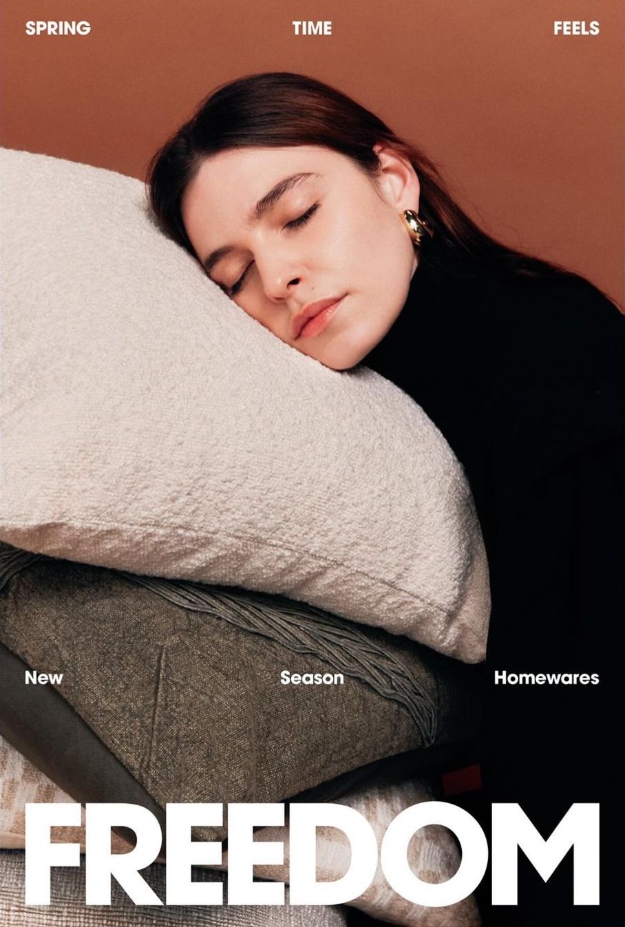 An adult woman rests her head on a stack of soft, neutral-toned cushions against a warm brown background, promoting home decor and relaxation.