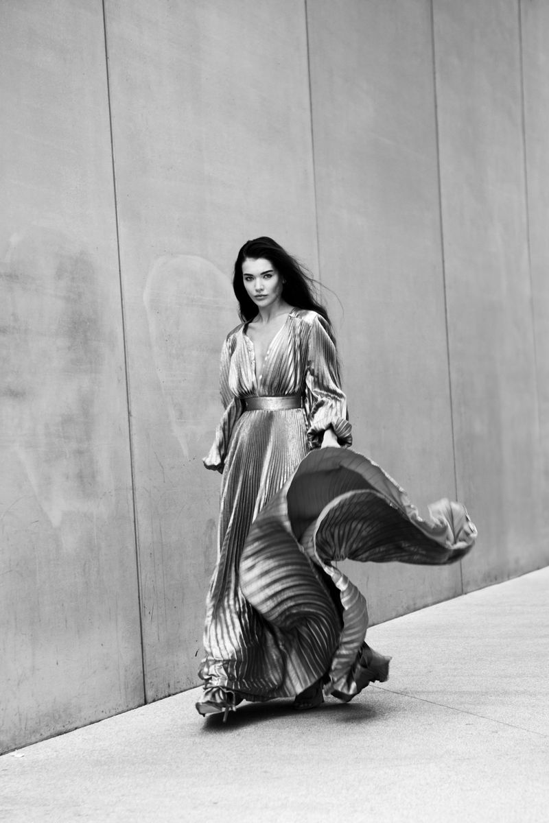 A woman in a pleated silver dress walks toward the camera on a sunny day, with concrete walls in the background. Fashion editorial.