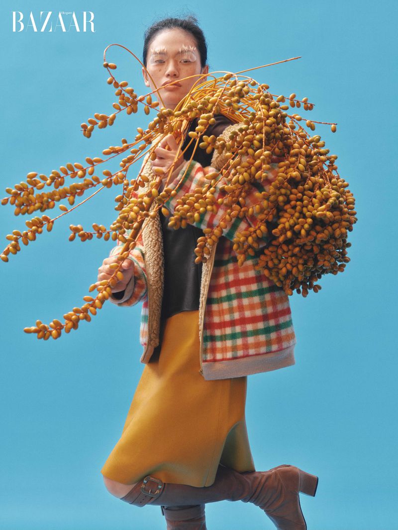 Fashion editorial shot: Woman in checkered jacket and boots poses with dried seed pods against a vibrant turquoise background.